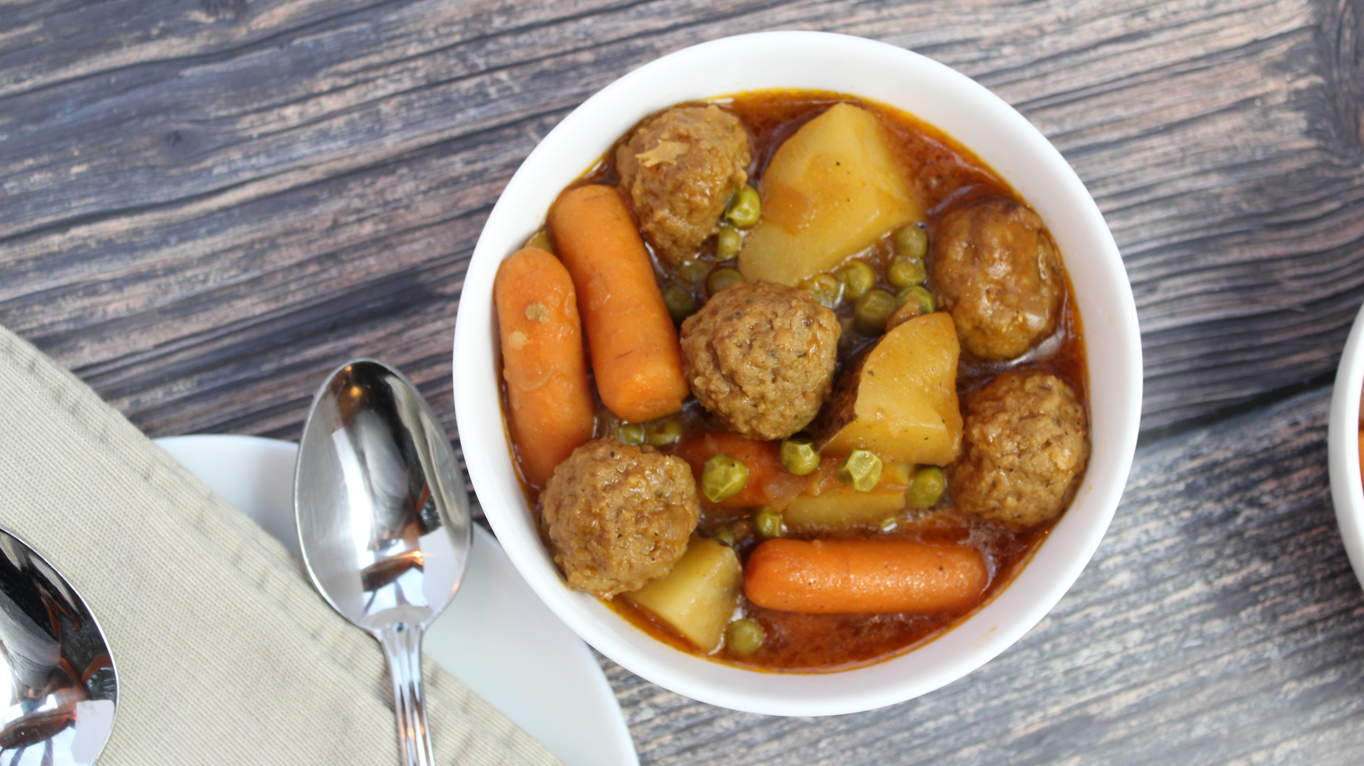 White bowl filled with meatballs, carrots, celery, potatoes and peas
