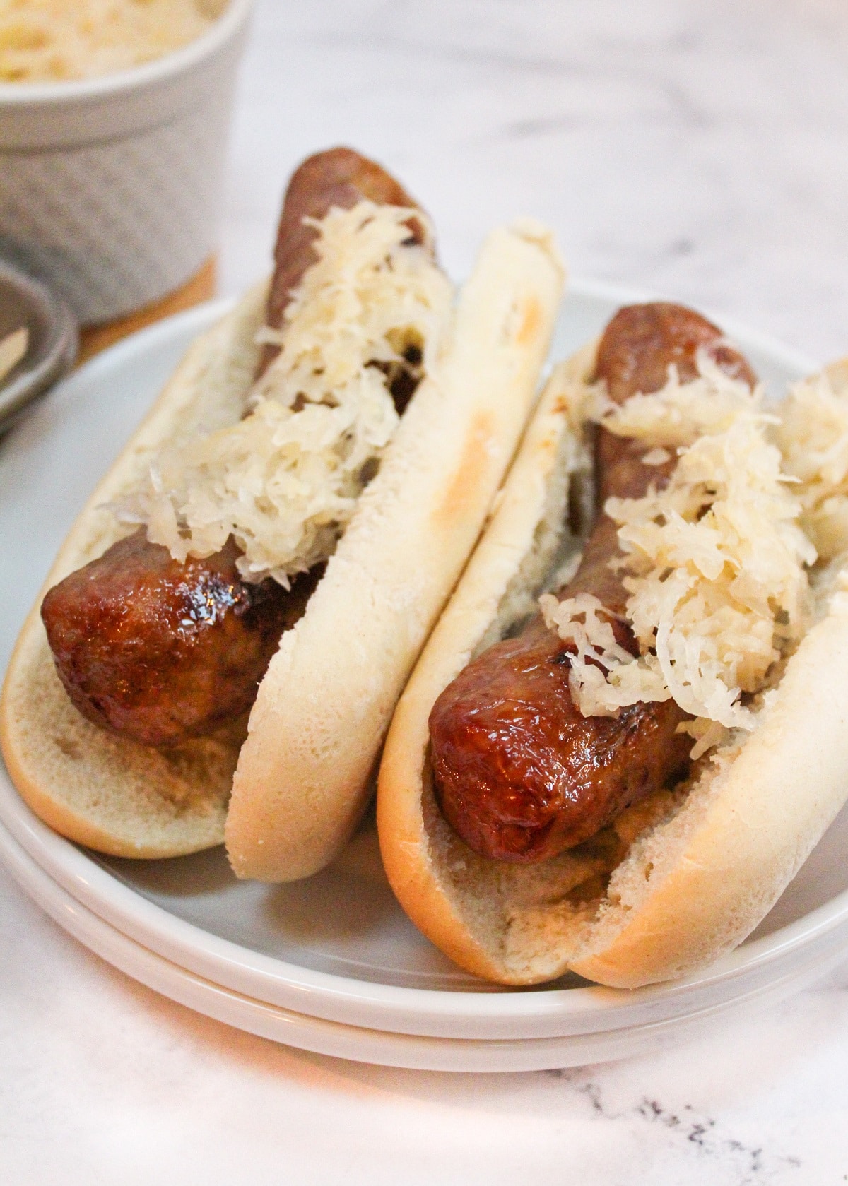 brats in a bun with sauerkraut on top on a white plate