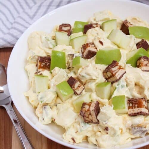 Creamy salad in a white bowl topped with chopped snickers bars and fresh green apple pieces.
