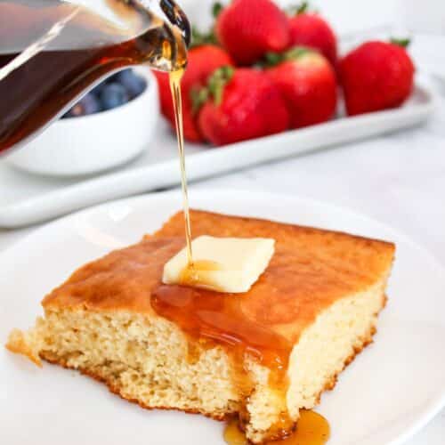 Plain pancake square on a white plate. Topped with butter. Maple syrup is poured from above over the pancake.
