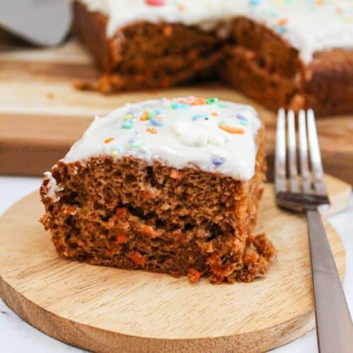Single slice of carrot cake on a wooden plate with a fork next to it. Topped with white frosting. The larger cake is in the background.